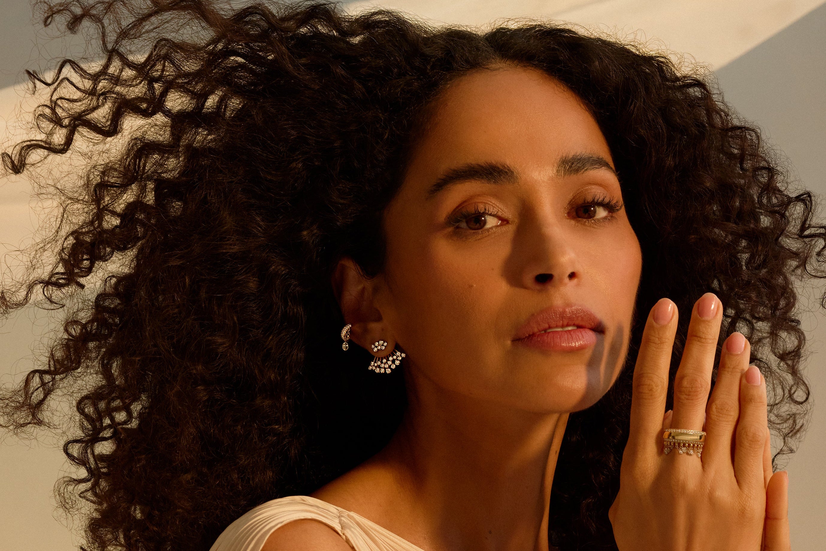 A woman with voluminous dark curly hair wearing a cream-colored pleated draped dress and Graziela Jewelry diamond silver earrings and ring, posing with her hands together in front of her face.